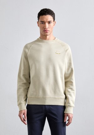 Lys beige genser, rund hals, ribbestrikkede ermer og hem, myk tekstur, har en liten gul logo på brystet, avslappet passform.
