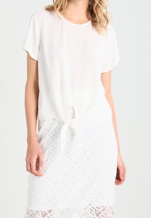 Femme portant un chemisier blanc à manches courtes avec un nœud à l'avant et une jupe blanche à motif de dentelle sur un fond uni.