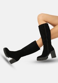 Botas altas de ante negras con un tacón grueso, cremallera lateral, suela de goma con un patrón de tracción, textura suave y diseño ajustado.