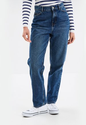 Persoon draagt een hoge taille blauwe spijkerbroek, witte sneakers en een wit shirt met lange mouwen met blauwe horizontale strepen.