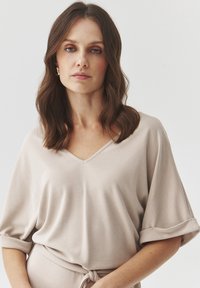 Top beige chiaro a V con maniche corte arrotolate, realizzato in un tessuto morbido. Presenta una vestibilità rilassata e un dettaglio con cintura annodata.