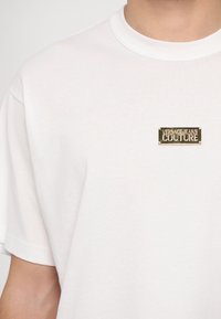 Baltas kokvilnas T-krekls ar apaļu dekoltē, kuram uz krūtīm ir zelta metāla logo plāksne ar uzrakstu "VERSACE JEANS COUTURE".