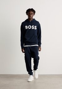 Hoodie azul marinho com o logótipo "BOSS", apresentando um bolso canguru, combinado com calças a condizer e ténis brancos. Textura de tecido suave, ajuste casual.
