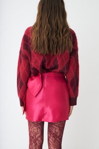 Czerwony i różowy sweter w wzory z motywem diamentów, w połączeniu z krótką, gorąco różową spódniczką z kokardą z tyłu i koronkowymi bordowymi rajstopami.