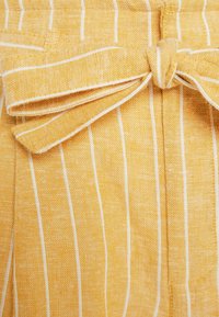 Keltainen raidallinen kangas, jossa on valkoisia pystysuoria viivoja. Vyötäröllä solmittava vyö, joka korostaa teksturoitua, kudottua ilmettä.