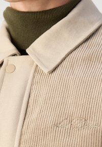 Veste en velours côtelé beige avec un col large, portée sur un pull à col roulé côtelé vert olive, texte brodé visible sur la veste.