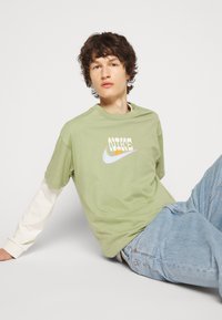 T-shirt en coton vert clair avec un imprimé graphique blanc et jaune sur le devant, superposé à une chemise blanche à manches longues. Jean en denim bleu clair.