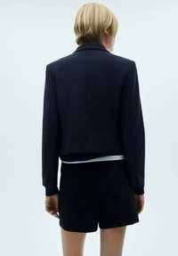 Navycroppierter Blazer mit langen Ärmeln, tailliertem Design und subtiler Textur, kombiniert mit schwarzen Shorts. Klare Linien und minimalistischer Stil.