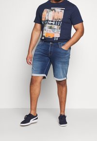 Muž v námořnickém tričku s grafikou surfovacích prken a oranžového vans, džínových šortkách s ohrnutým pasem a námořnických teniskách s bílou podrážkou.