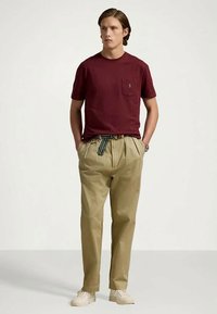 Ung man står med händerna i fickorna, klädd i en vinröd T-shirt, beige veckade byxor med randigt bälte, och vita sneakers mot en enfärgad bakgrund.