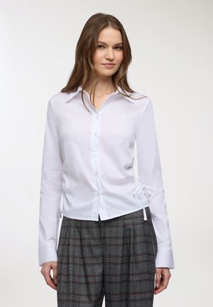 Giovane donna che indossa una camicia bianca attillata con bottoni e lacci laterali, accompagnata da pantaloni larghi grigi a scacchi su uno sfondo neutro.