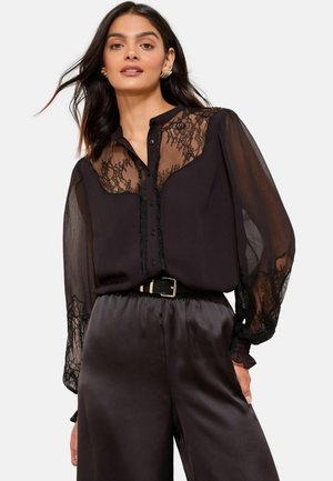 Schwarze Bluse mit durchsichtigem, spitzen Einsatz und langen, gepufften Ärmeln. Knopfleiste mit filigranen Spitzenverzierungen und Rüschendetails.
