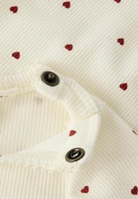 Biała tkanina z czerwonymi wzorami w serca; posiada napy wzdłuż krawędzi, podkreślające prążkowaną teksturę.