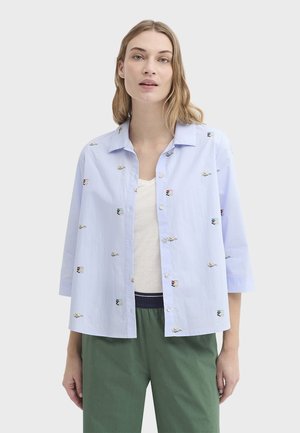 Svetlomodrá košeľa na gombíky s vyšívanými motívmi kníh, voľného strihu a s rukávmi do troch štvrtín. Nosená cez béžové tričko.