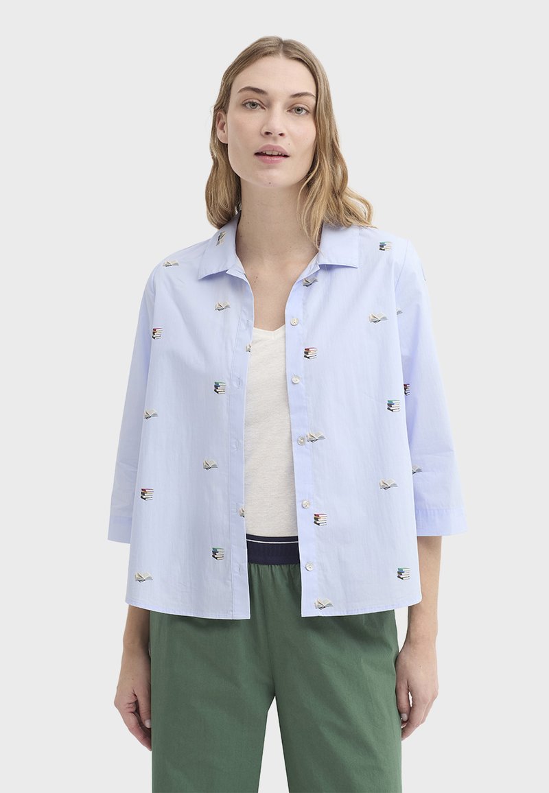 Chemise bleu clair boutonnée avec des motifs de livres brodés, coupe décontractée et manches trois-quarts. Portée par-dessus un haut beige.