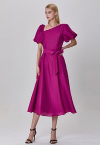 Robe fuchsia avec des manches bouffantes, un décolleté carré et un détail de taille à nouer, fabriquée dans un tissu lisse qui tombe jusqu'au mollet.