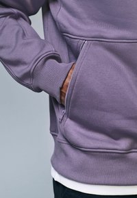 Felpa viola con tasca con zip, polsini e orlo a coste. Il materiale sembra morbido con una texture liscia e un design minimalista.