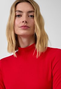Rode, langärmelige top met een hoge hals. Gladde textuur, aangesloten ontwerp. Model heeft schouderlang blond haar en neutrale make-up.