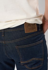 Jeans de mezclilla oscuros con costuras en color naranja y una etiqueta de cuero marrón en la parte trasera de la cinturilla. Dos bolsillos traseros visibles.