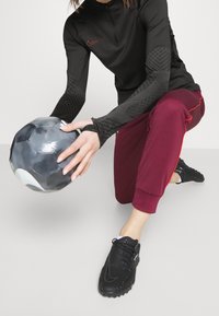 Schwarzes Sportoberteil mit halbem Reißverschluss und strukturierten Ärmeln, bordeauxrote, schmal zulaufende Jogginghose mit roten Akzenten und schwarzen Sportschuhen, hält einen schwarzen Fußball.