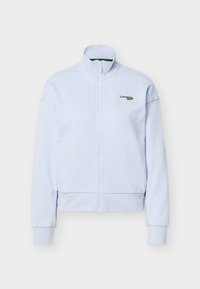 Veste zippée bleu clair avec col montant et manches longues, arborant un petit logo Lacoste vert sur la poitrine gauche.