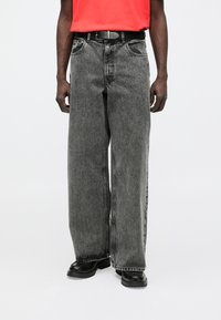 Jeans de perna larga em ganga preta desgastada com acabamento desbotado, com bolsos frontais e cinto preto, combinados com uma camisa vermelha brilhante.