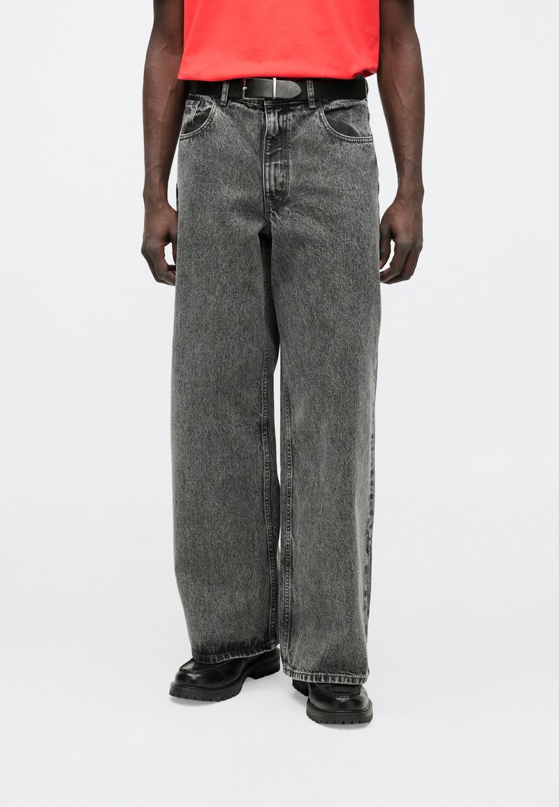 Τζιν με φαρδιά πόδια σε ξεθωριασμένο μαύρο denim με ξεβάμμα, διαθέτοντας μπροστινές τσέπες και μαύρη ζώνη, σε συνδυασμό με ένα φωτεινό κόκκινο πουκάμισο.