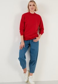 Sudadera de cuello redondo roja con mangas largas, combinada con unos vaqueros azules de corte amplio y zapatillas blancas. Tejido suave con puños acanalados.
