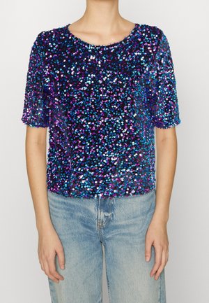 Kortærmet top dækket af blå, lilla og pink pailletter båret med lyseblå denimjeans på en person.