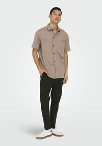 Camisa de botón de manga corta, color marr�ón claro, con dos bolsillos frontales, combinada con pantalones negros y zapatillas blancas con detalles en beige.