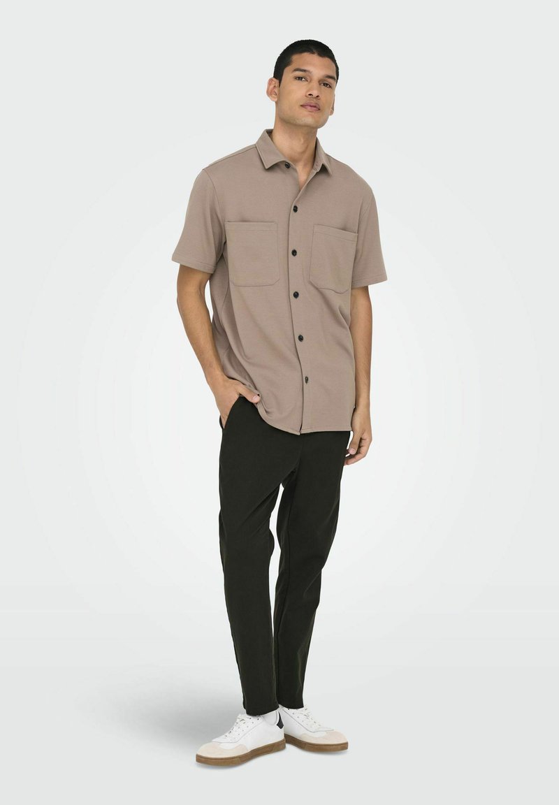 Camisa de botón de manga corta, color marrón claro, con dos bolsillos frontales, combinada con pantalones negros y zapatillas blancas con detalles en beige.