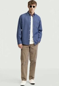 Blue and white striped button-up shirt, beige pants, white sneakers, and round sunglasses, worn by a model with a casual stance.