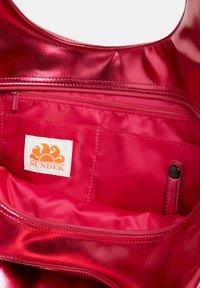 Interno di una borsa a tracolla metallica rossa con una tasca con zip, caratterizzata da un'etichetta bianca con un logo a forma di sole arancione. Texture liscia, design semplice.