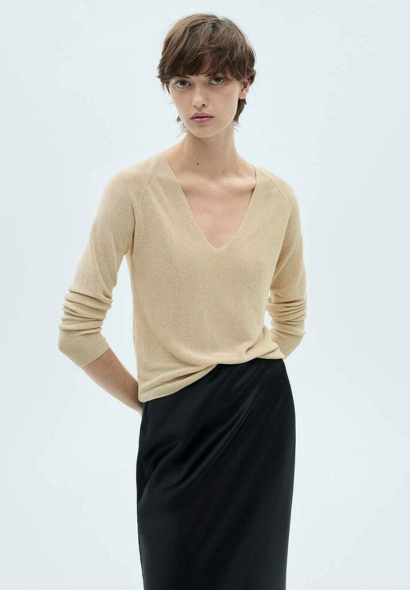 Beiger, langärmeliger Pullover mit V-Ausschnitt und strukturiertem Finish, kombiniert mit einem schwarzen, figurbetonten Rock, der eine schlichte, moderne Silhouette präsentiert.