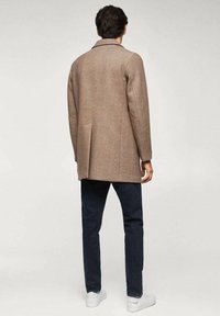 Brązowy płaszcz z mieszanki wełny o wyraźnej sylwetce, z pojedynczym rozcięciem i kołnierzem z wycięciem, zestawiony z ciemnymi dżinsami i białymi tenisówkami.