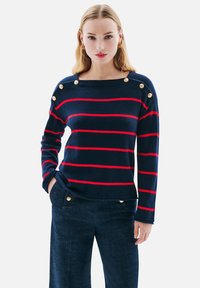 Pull marin à rayures avec des lignes horizontales rouges, col bateau, et détails en boutons dorés sur les épaules. Fait en tricot doux.
