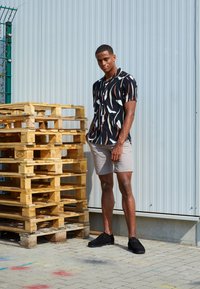 Chemise noire à manches courtes avec des motifs abstraits blancs et orange, associée à un short beige clair et des chaussures noires, à côté de palettes en bois.