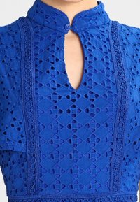 Vestido azul bordado con cuello alto y cierre de botón, recortes intrincados y un patrón texturizado en todo el vestido.