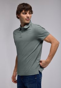 Polo de color verde claro hecho de algodón, con un collar clásico, tapeta de dos botones y mangas cortas. Combinado con jeans azules.