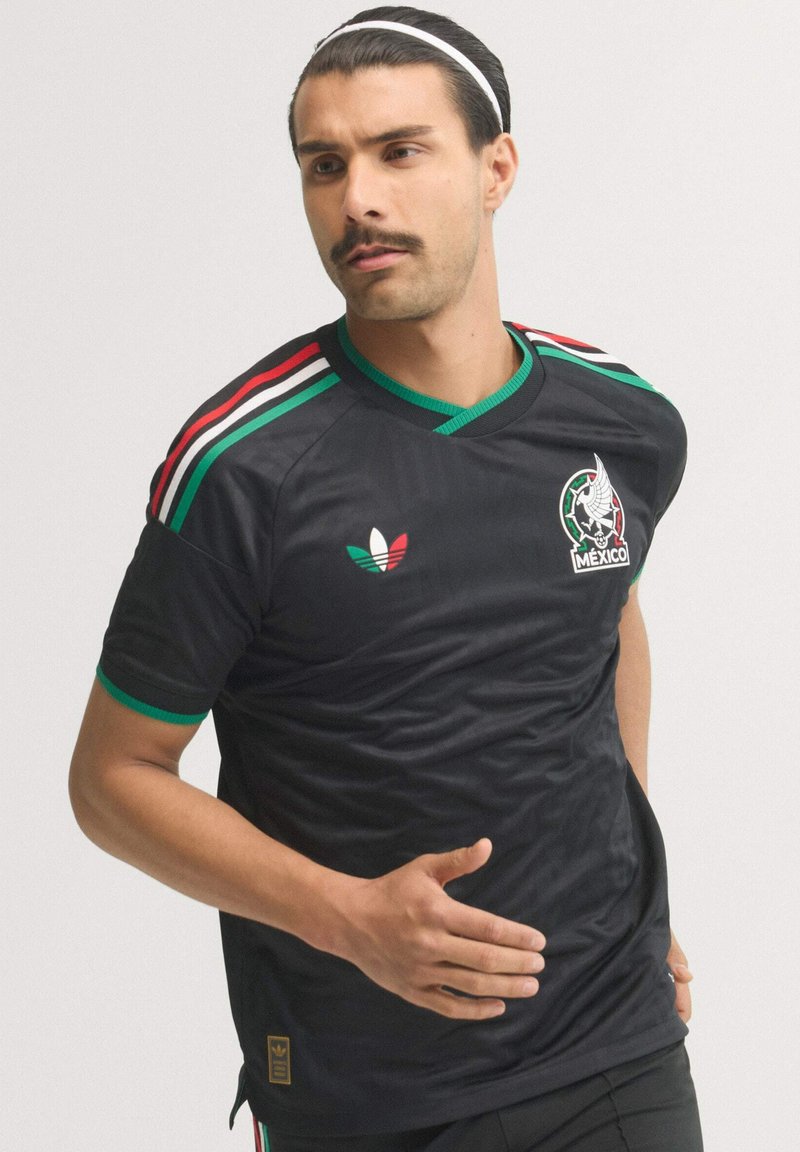 Man wearing black Mexico soccer jersey with red, white, green stripes on shoulders, a white headband, and posing with arm extended forward.