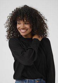 Black long-sleeve blouse with a smocked high neckline and puffed sleeves, paired with high-waisted denim jeans. Textured fabric, casual fit.
