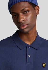 Marine polo-shirt med struktureret stof, to-knaps lukning og et gult ørnelogo på brystet. Strikket krave og korte ærmer.