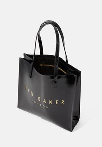 Sac en cuir verni noir avec deux poignées, arborant le logo doré "TED BAKER LONDON" sur le devant et une poche zippée à l'intérieur.