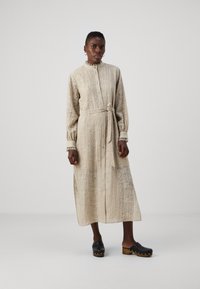 Long-sleeve, button-up dress in light beige fabric with a textured floral pattern. Includes a tie waist and cuff details. Worn with black clogs.