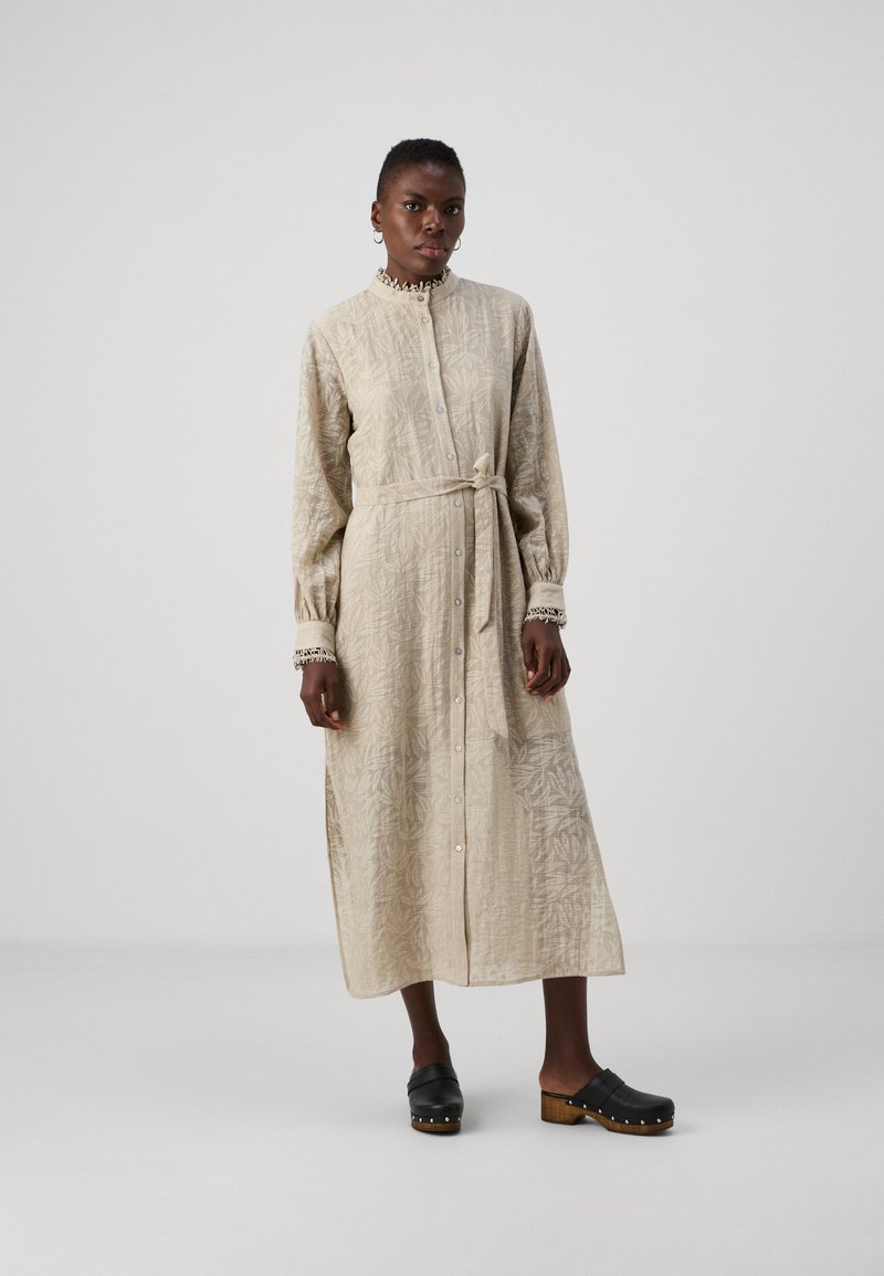 Long-sleeve, button-up dress in light beige fabric with a textured floral pattern. Includes a tie waist and cuff details. Worn with black clogs.