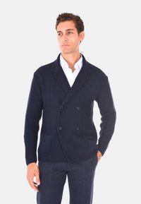 Cardigan in maglia doppiopetto blu navy con un motivo testurizzato, due tasche frontali e un colletto a scialle. Indossato sopra una camicia bianca.