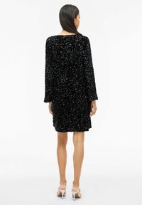 Juoda blizgi suknelė su ilgomis rankovėmis, iki kelių ilgio kirpimu ir laisvo silueto. Lygios audinio tekstūros, su apvalia iškirpte.
