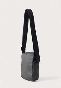 Pieni suorakulmainen musta olkalaukku, jossa leveä säädettävä hihna, sileä kangas ja minimalistinen muotoilu yksinkertaisella taustalla.