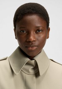 Beige Trenchcoat mit großem, gefaltetem Kragen und geknöpften Schulterklappen, aus einem glatten, strukturierten Stoff und mit klaren Linien.