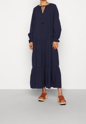 Kvinde iført en lang navy lagdelt kjole med lange puffede ærmer og brune åbne træsandaler med platform, stående på gråt gulv.